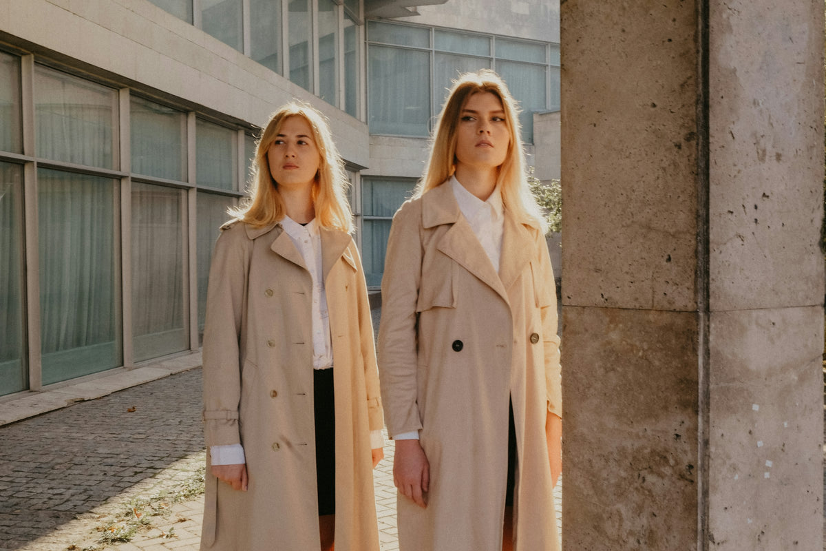 Two women in camel trench coats outside a stone building